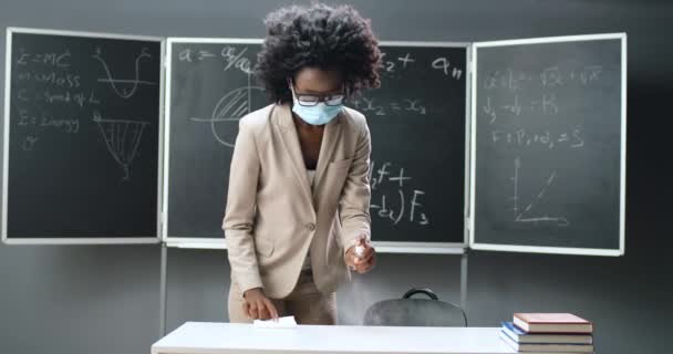 Fiatal afro-amerikai tanárnő szemüvegben és orvosi maszkban előkészíti az osztálytermet és az asztalt a leckéhez. Női permetező fertőtlenítő az asztalon, és tisztítsa meg a baktériumok. Coronavirus pandémia. — Stock videók