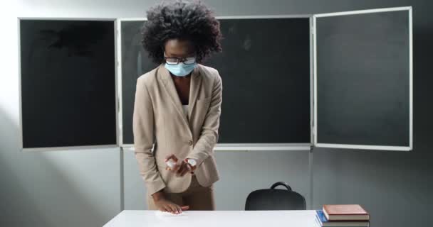 Femme afro-américaine enseignante en lunettes et masque médical préparant salle de classe et table pour la leçon. Désinfectant de pulvérisation femelle sur le bureau et désinfectant des germes. Pandémie de Covid-19. Vie scolaire. — Video