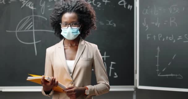 Πορτρέτο της νεαρής Αφρο-Αμερικανίδας δασκάλας με γυαλιά και ιατρική μάσκα κοιτάζοντας την κάμερα στην τάξη και κρατώντας σημειωματάρια. Μαύρη σανίδα με φόρμουλες στο παρασκήνιο. Πανδημία. — Αρχείο Βίντεο