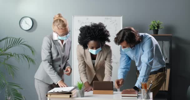 Tres razas mixtas de hombres y mujeres con máscara médica de pie en la oficina y viendo algo en el teléfono inteligente. Profesores multiétnicos discuten el concepto en línea de estudiar por teléfono móvil. Hombres y mujeres. — Vídeo de stock