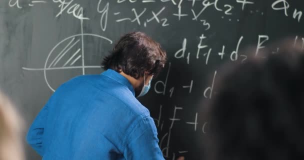 Caucasian man teacher in medical mask at school writing formulas and mathematics laws on blackboard. School concept. Male lecturer in glasses explaining physics laws. Coronavirus concept. — Stock Video