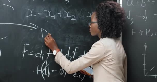 黒板に数式や数学の法律を書く学校でアフリカ系アメリカ人の若い女性教師についてのバックビュー。学校のコンセプト。物理学の法則を説明する眼鏡の女性講師。リア. — ストック動画