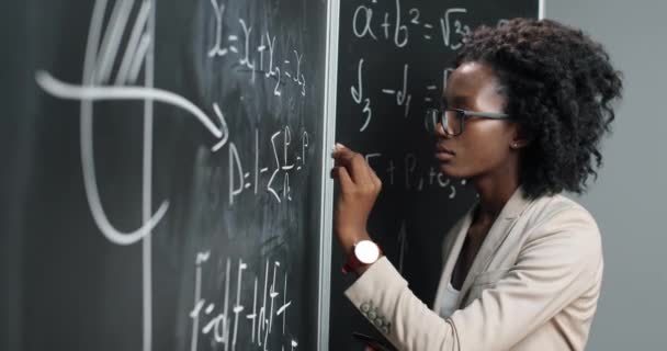 Profesora joven afroamericana en la escuela escribiendo fórmulas y leyes matemáticas en pizarra. Concepto escolar. Profesora de matemáticas en gafas explicando leyes de física. Educación. — Vídeos de Stock