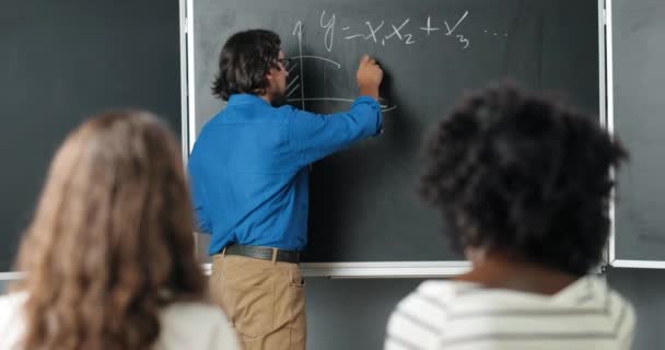Vit manslärare på skolan skriver formler och matematik lagar på svarta tavlan. Skolbegreppet. Manlig föreläsare i glasögon som förklarar matematiska lagar för eleverna. Utbildningsidé. — Stockvideo