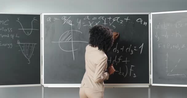 Hátul az afro-amerikai fiatal matematika vagy fizika tanárnő képleteket ír táblára krétával. Iskolai tanár nő fordult kamera és elmagyarázza a matematika. Visszanézés. — Stock videók