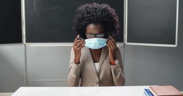 Porträtt av afroamerikansk kvinnlig lärare i glasögon som tar på sig medicinsk mask, sitter vid bordet i klassrummet i skolan och ler mot kameran. Kvinnoföreläsare som bär skydd. Begreppet coronavirus. — Stockvideo