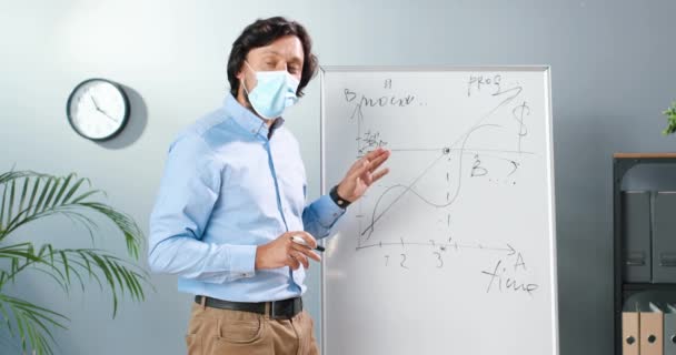 Caucasian man teacher in medical mask and glasses standing at board in classroom and telling physics or geometry laws to class. Coronavirus concept. School during covid-19. Educative lection. — Stock Video