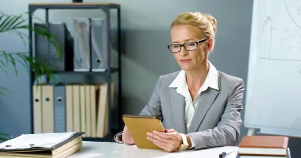照片上的白种人女商人戴着眼镜坐在办公室里，使用平板设备，对着相机微笑。女性在橱柜桌上的小玩艺儿电脑上的商务风格的敲击和滚动. — 图库视频影像