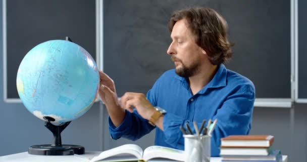 Retrato de un maestro caucásico sentado en la mesa y examinando el globo en clase. Hombre enseñando geografía en el aula. Lección en línea. Profesor explicando el tema del mundo. — Vídeos de Stock