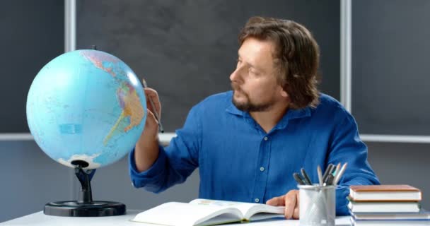 Portret białego nauczyciela siedzącego przy stole i badającego globus w klasie, notujący w podręczniku ołówkiem. Człowiek czytający książkę o geografii w klasie. Lekcja online. Wykładowca przygotowujący się do odprawy — Wideo stockowe