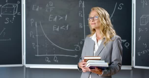 Potret guru wanita Kaukasia yang cantik berdiri di dalam kelas, tersenyum kepada kamera dan memegang buku pelajaran. Dosen wanita dengan buku di papan tulis dengan rumus matematika dan gambar. — Stok Video