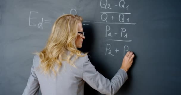 Vue arrière sur conférencière caucasienne écrivant des formules mathématiques ou physiques avec des craies sur le tableau noir. Portrair d'une enseignante travaillant à l'école et souriant à la caméra. Leçon de mathématique. Arrière. — Video