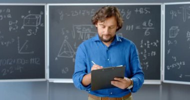Sınıfta duran beyaz erkek öğretmenin portresi. Ellerindeki klasöre notlar yazıyor. Matematik formülleri ve arka planda yasalar. Erkek matematikçi, okul ya da üniversitede çalışıyor..