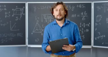 Sınıfta elinde klasörle kameraya bakan beyaz erkek öğretmenin portresi. Matematik formülleri ve arka planda yasalar. Erkek matematikçi, okul ya da üniversitede çalışıyor..