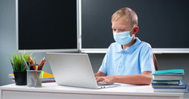 Tıp maskeli küçük beyaz çocuk okulda masasında oturuyor. Ödev yapan ve dizüstü bilgisayarda yazan bir öğrenci. Coronavirus salgını konsepti. Öğrenci okulda okuyor..