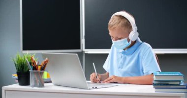 Tıp maskeli ve kulaklıklı küçük beyaz çocuk okulda masada oturuyor. Ödev yapan ve bilgisayarın önünde defter kitabı yazan genç bir öğrenci. Coronavirüs salgını konsepti.