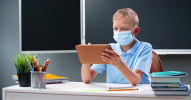 Tıp maskeli küçük beyaz bir çocuk okulda masada oturuyor ve tablet cihazıyla oynuyor, ekranda tıkırdıyor ve kayıyor. Küçük okul çocuğu alet bilgisayarı ve mesajlaşma kullanıyor. Salgın konsepti.