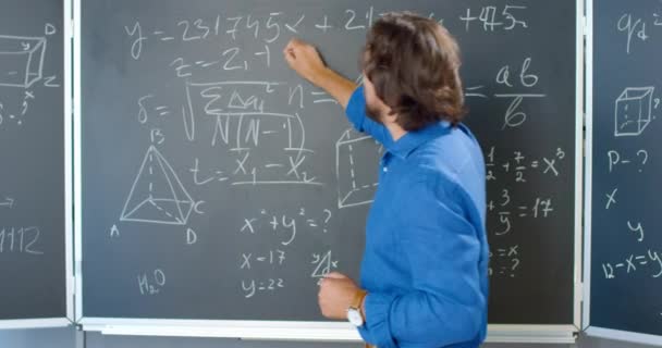 Professeur masculin caucasien debout au tableau noir avec des formules, des graphiques et des lois et expliquant les mathématiques. Un homme écrit à bord avec de la craie. Professeur de mathématiques à l'école ou au collège. — Video