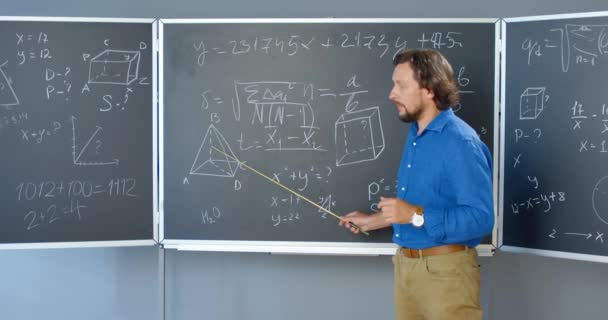 Caucasian handsome male teacher standing at blackboard with formulas, graphics and laws and explaining with pointer. Lecturer of mathematics at school or college. Math lection. — Stock Video