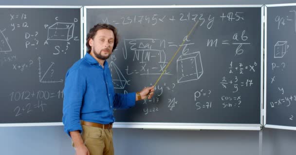 Fehér férfi matektanár áll a táblánál képletekkel, grafikákkal és törvényekkel, és magyarázza a fizikát mutatóval. Matematika tanár az iskolában vagy a főiskolán. Tanítási koncepció. — Stock videók
