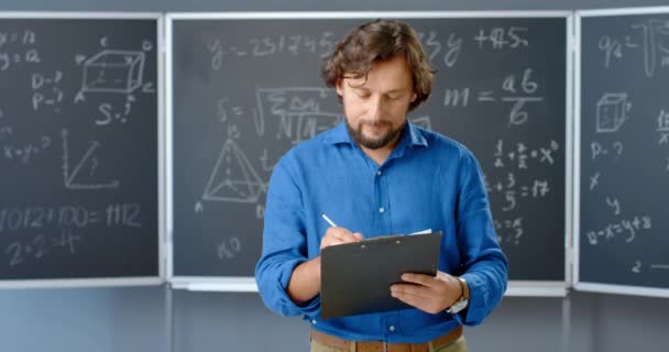 Portret van een blanke mannelijke leraar die in de klas staat en notities schrijft op papier in mappen in handen. Wiskunde formules en wetten op de achtergrond. Docent wiskunde op school of op de universiteit. — Stockvideo