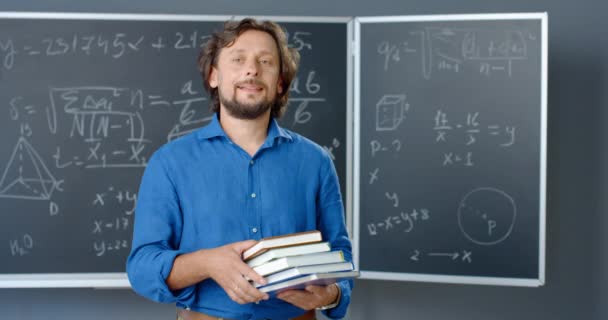 Retrato de un maestro caucásico de pie en el aula con libros en las manos mirando a la cámara. Fórmulas matemáticas y leyes de fondo. Hombre matemático sosteniendo libros de texto que trabajan en la escuela o la universidad. — Vídeos de Stock