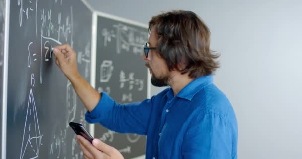 Großaufnahme eines kaukasischen männlichen Lehrers mit Brille, der Formeln und Mathegesetze auf die Tafel schreibt und aufs Smartphone schaut. Lehrstunde in Mathematik. Mann benutzte Handy als Schreiberling. — Stockvideo