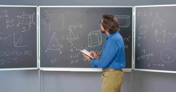 백보드에 안경을 쓴 백인 남자 수학자 수학이나 식물 공식, 그리고 노트북에 노트를 작성하는 법률. 학교에서 교사가 교과서로 대수학이나 기하학 방정식을 확인하는 모습. — 비디오