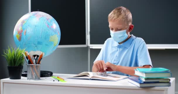 Pequeño chico adolescente caucásico en máscara médica sentado en el escritorio con el mundo en la escuela. Adolescente escolar leyendo libro de texto y volteando páginas. Aprendizaje de los alumnos en el aula. Concepto de Coronavirus. — Vídeos de Stock