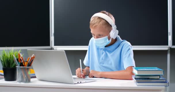 Pieni valkoihoinen teinipoika lääketieteellisessä naamiossa ja kuulokkeet pöydällä koulussa. Teini-ikäinen koulupoika tekee läksyjä ja kirjoittaa liikuntaa copybookissa kannettavan tietokoneen edessä. Covid-19 konsepti. — kuvapankkivideo