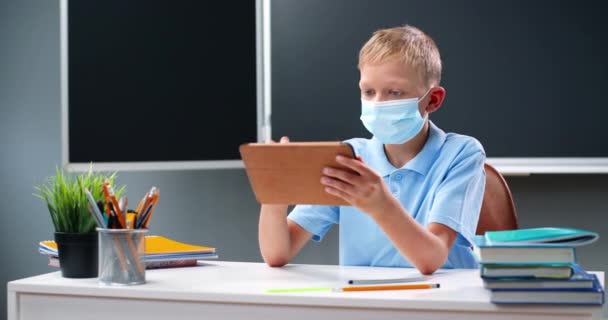 Caucasiano pequeno adolescente em máscara médica sentado na mesa na escola e jogando no dispositivo tablet, tocando e rolando na tela. Pequeno estudante usando o computador gadget e mensagens de texto. Conceito de pandemia. — Vídeo de Stock