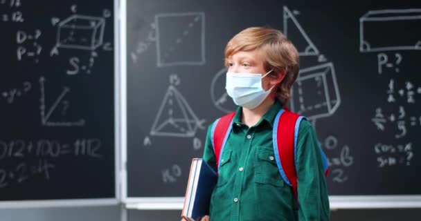 Portrait of cute red-haired small Caucasian boy in medical mask and with backpack standing in classroom, turning to camera and holding textbooks. Little schoolboy with red hair in school. Covid-19. — Stock Video