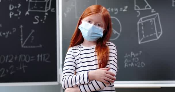 Portrait de jolie petite fille aux cheveux roux caucasienne avec sac à dos et masque médical debout dans la salle de classe et se tournant vers la caméra. Écolière rousse à l'école au tableau noir. Pandémie de coronavirus — Video