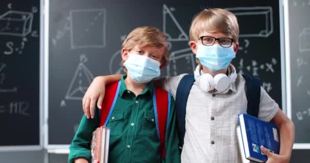 Porträtt av söta små kaukasiska pojkar bästa vänner i medicinska masker, med ryggsäckar och läroböcker står i klassrummet vid svarta tavlan. Små skolpojkar kramas i skolan. Coronaviruspandemi. — Stockvideo