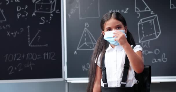 Портрет милої щасливої маленької кавказької дівчини з рюкзаком, що стоїть у класі, дивиться на камеру і знімає медичну маску. Маленька іспанська школярка посміхається в школі. Коронавірус. — стокове відео