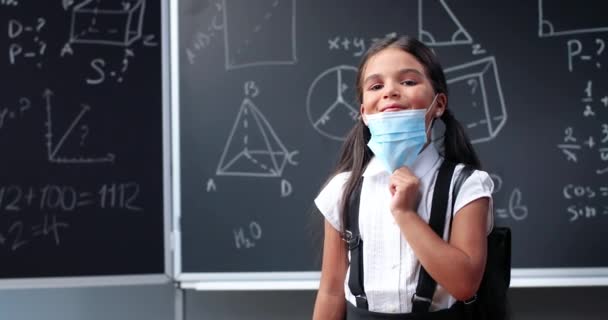 Portret van een leuk klein Latino meisje met rugzak in de klas, lachend naar de camera en zonder medisch masker. Klein Spaans schoolmeisje op schoolbord op school. Coronaviruspandemie. — Stockvideo