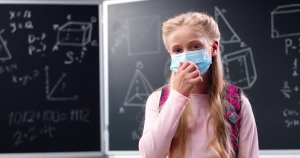 Портрет досить щасливої дівчинки з кавказьким рюкзаком, що стоїть у класі, дивиться на камеру і знімає медичну маску. У школі дівчина посміхається на дошці. Pandemic Covid-19. — стокове відео