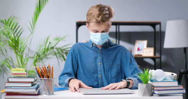 Teenager běloch v lékařské masce sedí za stolem ve škole. Teenage školák dělat domácí úkoly a psaní na notebooku. Koronavirová pandemická studie. Žák, který studuje ve škole. — Stock video