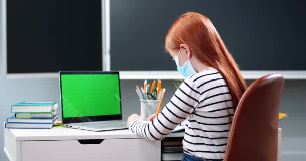 Portret pięknej białej dziewczyny z długimi czerwonymi włosami i maską medyczną piszącej pracę domową w copybooku, patrzącej w kamerę i siedzącej przy biurku. Laptop komputer z zielonym ekranem. Klucz chromowy. — Wideo stockowe