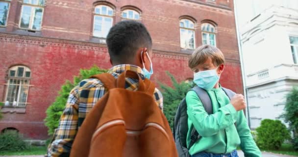 Ritratto ravvicinato di bambini multietnici in maschere mediche che salutano con gomiti vicino a scuola. Ragazzi afroamericani e caucasici carini in piedi all'aperto con gli zaini. Vita degli alunni Concetto di quarantena — Video Stock