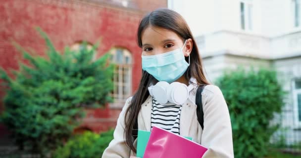 Κοντινό πορτραίτο ενός όμορφου καυκάσιου κοριτσιού με ιατρική μάσκα μπροστά από το σχολείο. Χαριτωμένο σχολιαρόπαιδο που στέκεται με ακουστικά και βιβλία σε καραντίνα. Έννοια μαθητών — Αρχείο Βίντεο