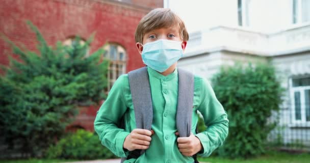 Κοντινό πορτραίτο ενός χαριτωμένου καυκάσιου αγοριού με ιατρική μάσκα μπροστά από το σχολείο. Μαθητής με σχολική τσάντα σε καραντίνα. Μελέτη κατά τη διάρκεια πανδημίας. Έννοια ιού — Αρχείο Βίντεο