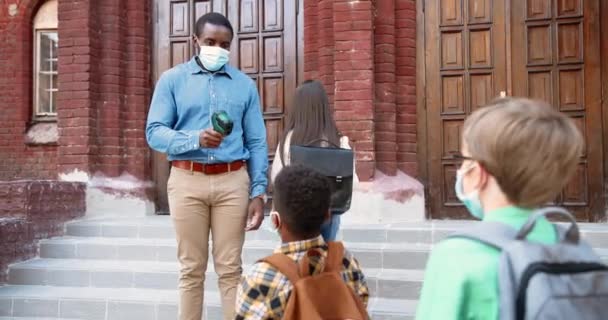 African American handsome male teacher measuring temperature of multi-ethnic pupils with infrared thermometer in front of school. After quarantine study. Healthcare control Preventive measures concept — Stock Video