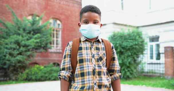 Портрет милого счастливого маленького афроамериканского мальчика в медицинской маске на школьном дворе. Школьник-младший, стоящий с сумкой на улице после карантина. Учеба во время пандемии. Концепция учащихся — стоковое видео