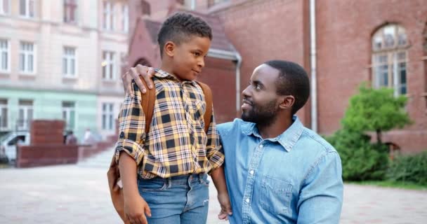 Portrét veselého afroamerického dospívajícího chlapce stojícího se šťastným pohledným otcem poblíž školy. Radostný muž s roztomilým synem s batohem s úsměvem na kameru venku. Zpátky do školy. Rodinný koncept — Stock video