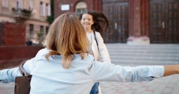 Zamknij portret szczęśliwej dziewczynki biegnącej do matki po zajęciach w szkole. Tył białej kobiety spotyka córkę ze szkoły na świeżym powietrzu i przytula się w dobrym nastroju. Koncepcja rodziny — Wideo stockowe