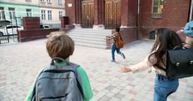 Sırt çantalı, dışarıda okula koşan mutlu çoklu etnik çocuklar. Afro-Amerikalı oğlan ve beyaz kız defterleri ile sınıflara koşuyor. Erkek ve bayan öğretmenler arka planda okulun yanında duruyorlar.