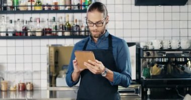 Bardaklı ve önlüklü beyaz bir erkek mutfaktaki kafede tablet kullanıyor. Bar 'daki yakışıklı garson tablet bilgisayarda internete bağlanıp kayıyor. Gadget kullanıcısı. Yakınlaştırılıyor.