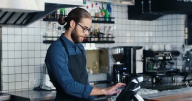 Genç yakışıklı beyaz barmen tezgahta duruyor ve bardaki bilgisayara düzen getiriyor. Barmen çalışıyor ve faturaları sayıyor. Gözlüklü ve önlüklü adam..