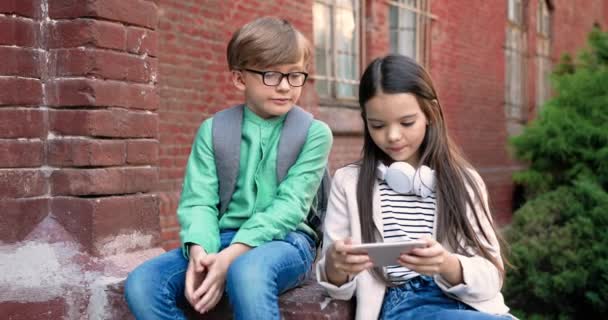 Portret nastolatków rasy kaukaskiej siedzących na świeżym powietrzu koło szkoły. Ładna dziewczyna pisząca i stukająca na smartfonie i rozmawiająca z przyjacielem. Chłopiec w okularach z plecakiem patrząc na ekran telefonu — Wideo stockowe